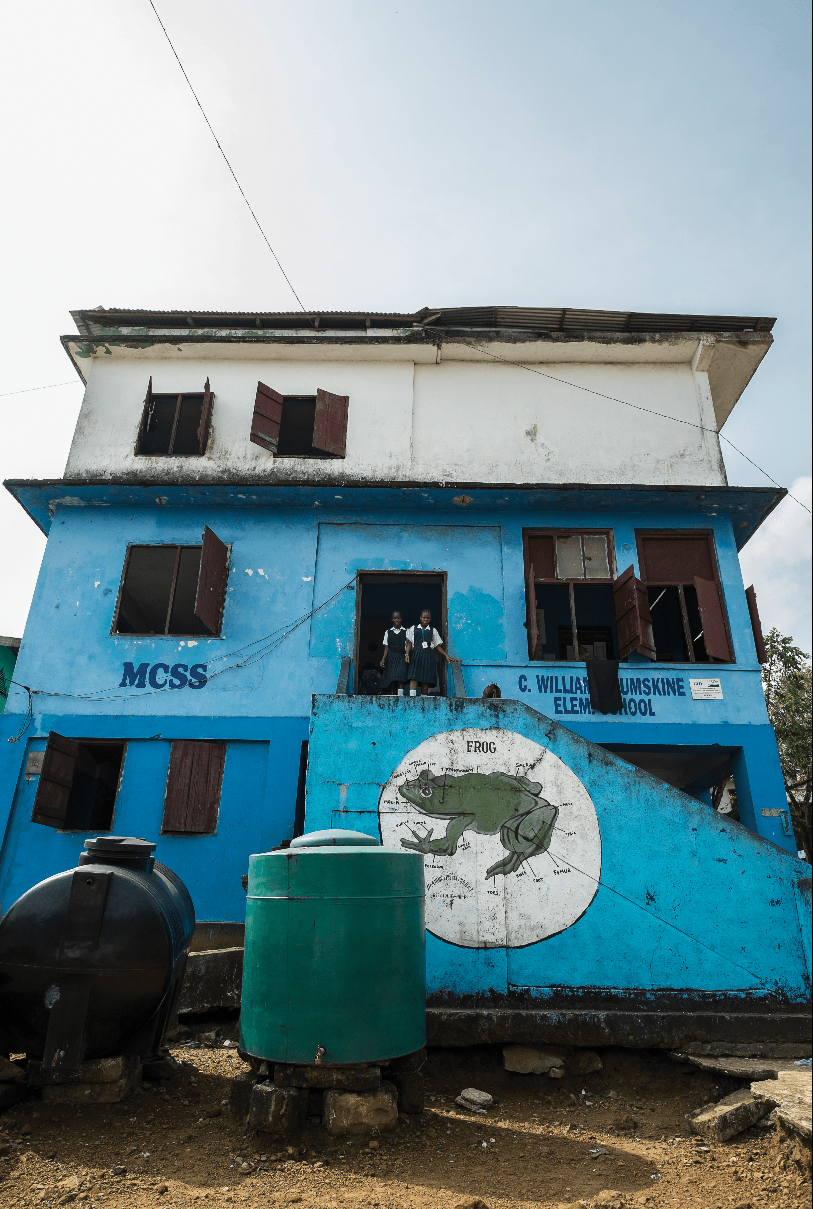 Photo of 3-storey blue Liberian school with two students in doorway